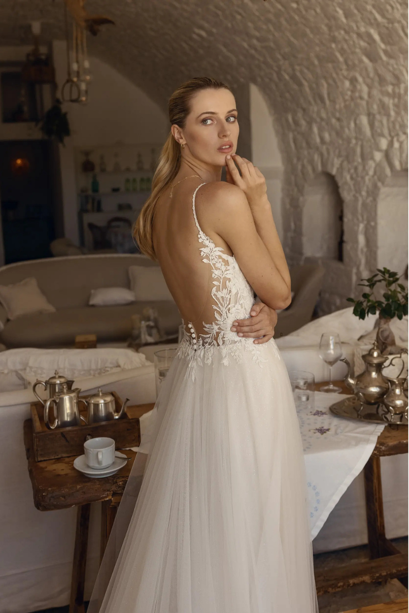 Eine Frau in einem weißen Brautkleid namens Alinde steht mit leicht zur Seite gedrehtem Oberkörper und blickt in die Kamera. Das Kleid hat einen tiefen Rückenausschnitt und ist mit Spitze und floralen Applikationen verziert. Ihre Hand ist an ihrem Kinn. Sie steht in einem hellen Raum mit Steinwänden und einem Tisch mit silbernem Geschirr.