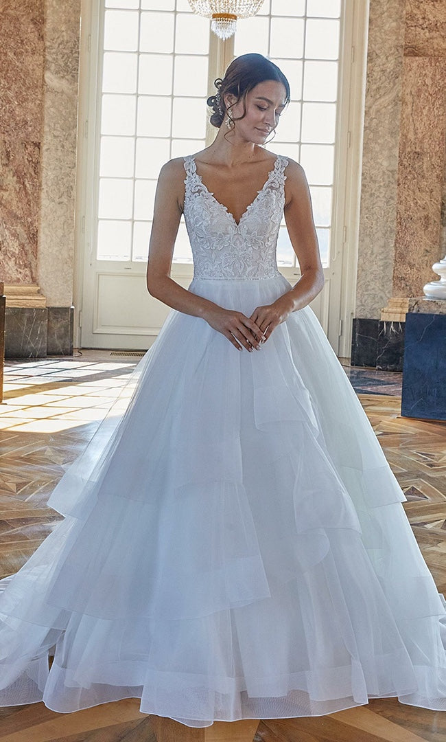 Celestia“ A-Linien-Brautkleid von „Susannas Brautmode“, mit einem tiefen V-Ausschnitt und einem mehrlagigen Tüllrock, präsentiert in einem eleganten Raum, ideal für die stilvolle Rheinland-Braut.
