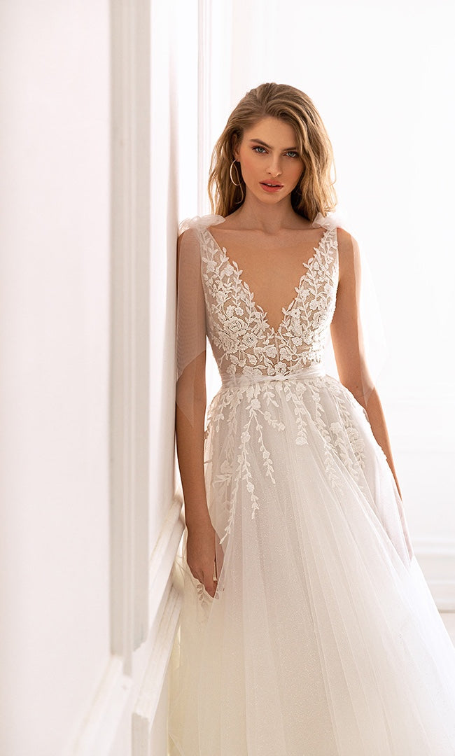 Braut in einem A-Linien-Hochzeitskleid mit V-Ausschnitt und Spitzenoberteil, umrahmt von zartem Tüll, steht in einem hell erleuchteten Raum.