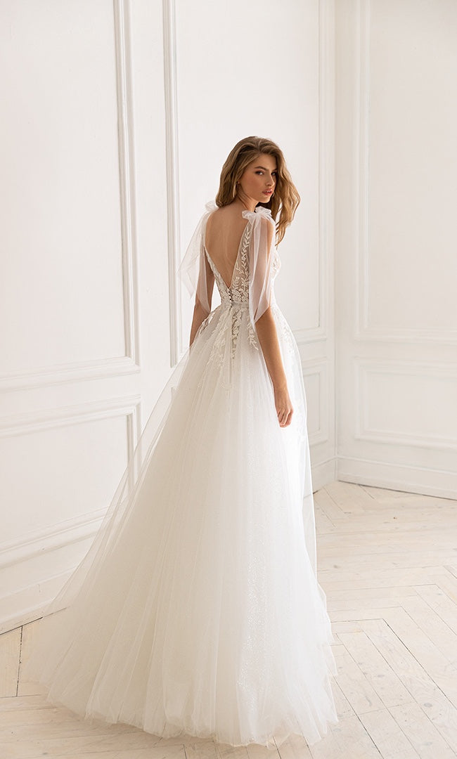 Braut steht in einem A-Linien-Hochzeitskleid mit tiefem V-Rückenausschnitt und Spitzenapplikationen, die den Rücken betonen, vor einem lichtdurchfluteten Hintergrund.