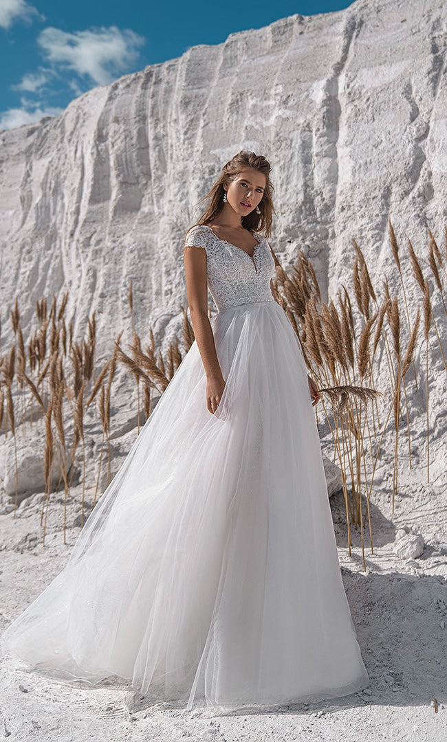 Braut in "Lumina" A-Linien-Brautkleid von "Susannas Brautmode" mit feiner Spitze und einem majestätischen Tüllrock vor einer strahlend weißen Kalkwand, das den Glanz einer Hochzeit im Rheinland widerspiegelt.