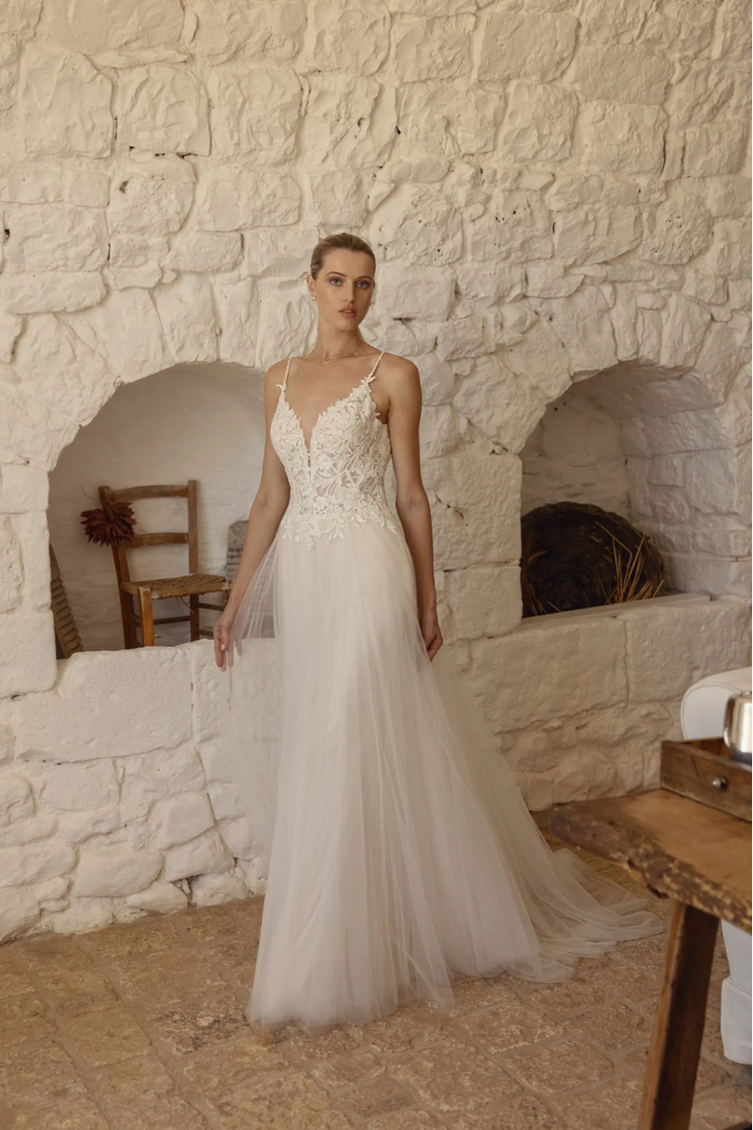  Eine Frau in einem bodenlangen, weißen Brautkleid namens Alinde steht aufrecht in einem Raum mit einer rustikalen Steinwand. Das Kleid hat Spaghetti-Träger, einen tiefen V-Ausschnitt und ist mit Spitze am Oberteil verziert, der Rock fällt weit und fließend herab.