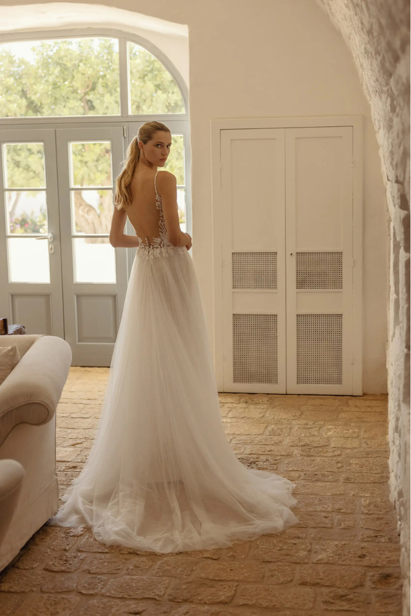 Eine Frau in einem langen, weißen Brautkleid namens Alinde steht mit dem Rücken zur Kamera und blickt über ihre Schulter nach hinten. Das Kleid hat einen tiefen Rückenausschnitt und einen weiten Tüllrock. Sie steht in einem Raum mit hellen Steinmauern und großen Fenstertüren, die ins Freie führen.
